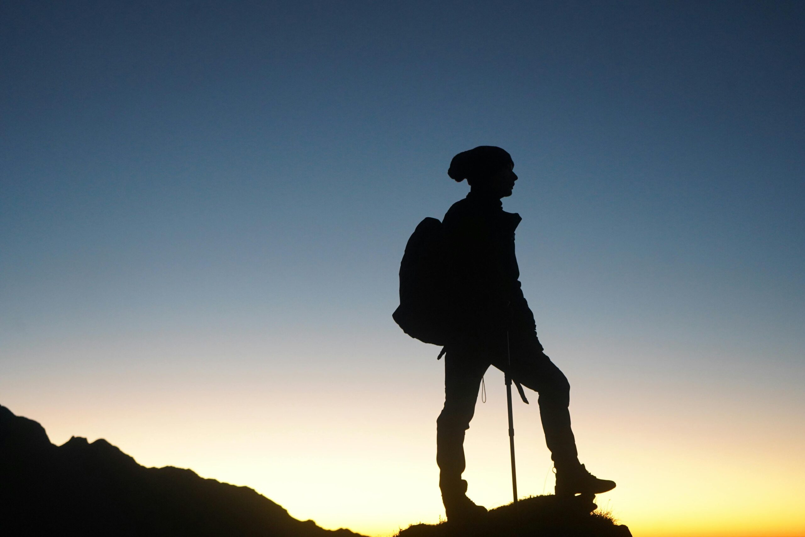 Person standing on top of a rock, wearing a backpack and holding a walking cane, against the backdrop of a sunset