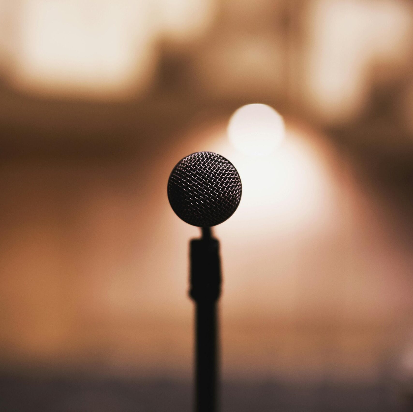 Image of a microphone in front with in the background a faded theatre in beige and brown colours including spotlight