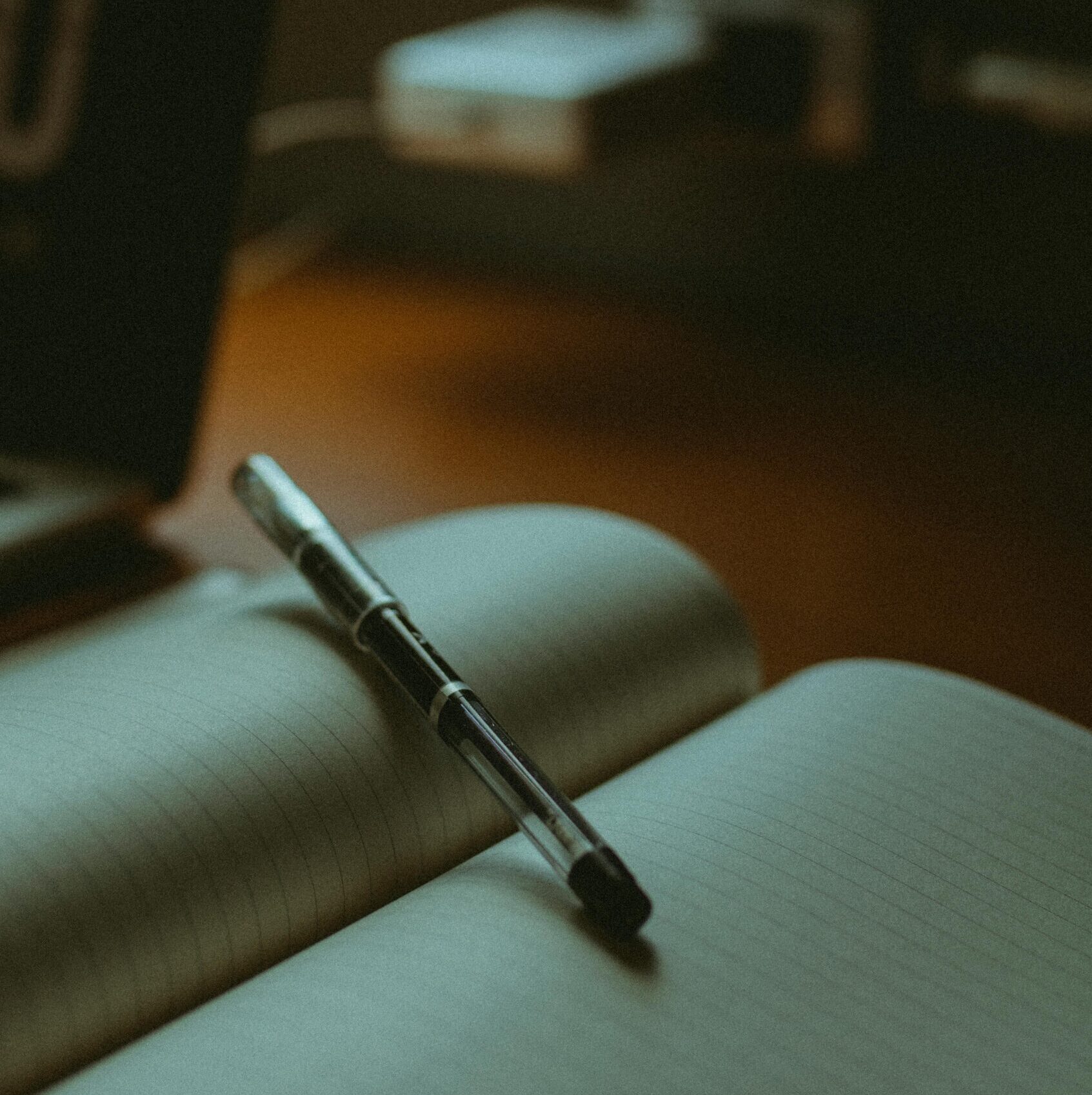 Image of a crowded desk space, zoomed in on an open notebook with a pen balancing on its pages