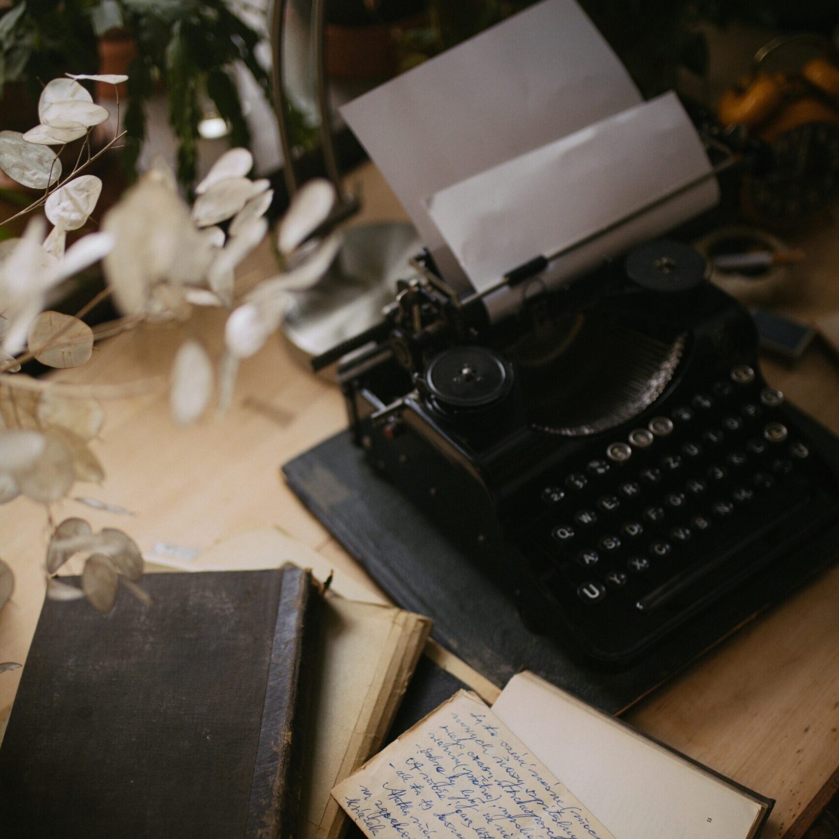 Old typewriter standing on a desk made of wood, with lots of notebooks and scribbles next to it and multiple plants around it
